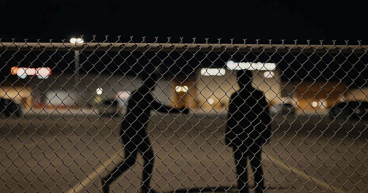 Chain-link fence at night separating silhouetted figures in a venue parking lot with blurred lights behind