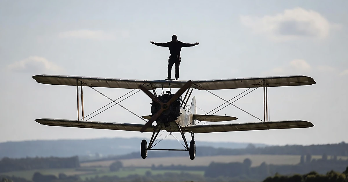 Biplane in flight with wing walker silhouette strapped to upper wing against bright sky at airfield