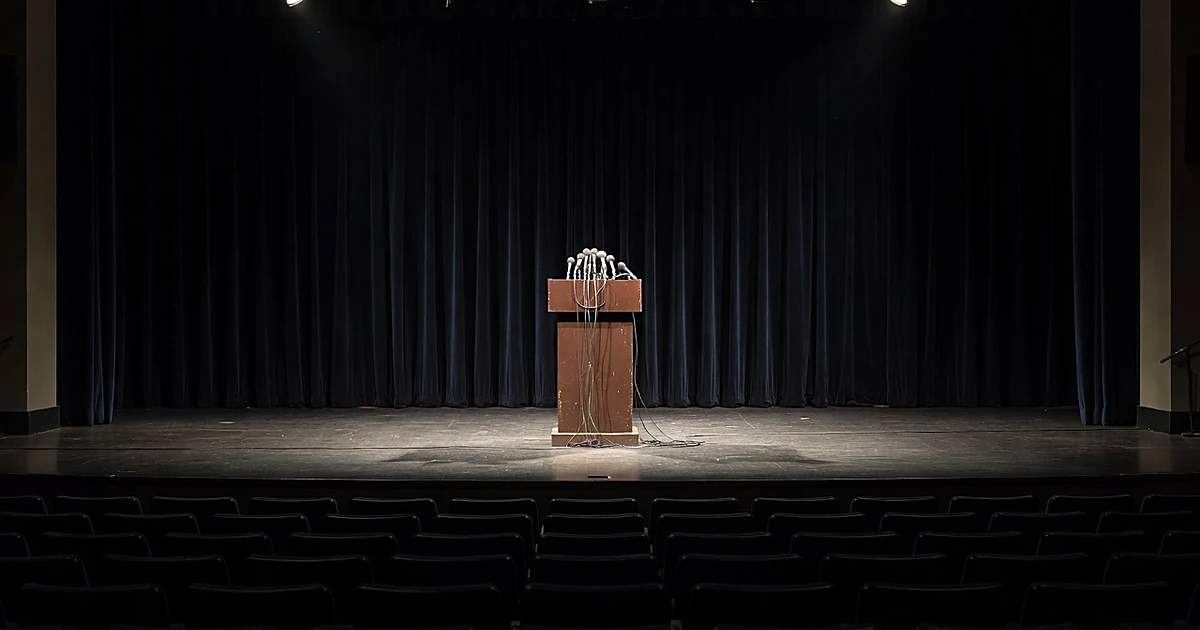 Empty debate podium with microphones under stage lights, surrounded by vacant chairs in darkened auditorium
