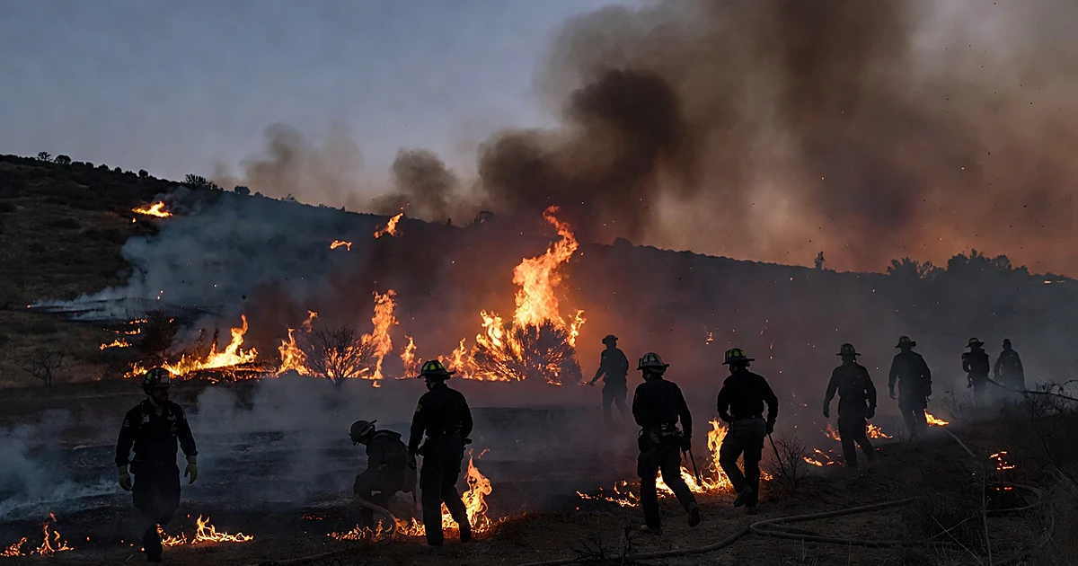 Wildfire flames consuming hillside brush at dusk with firefighter silhouettes in smoky foreground