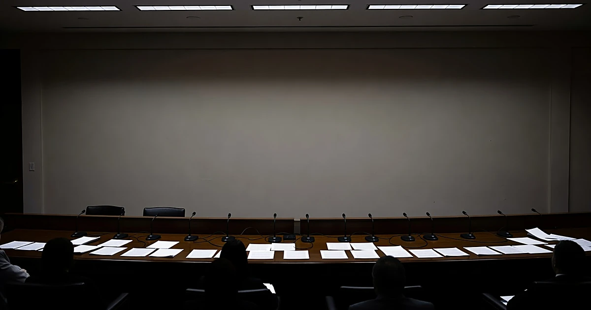 Congressional hearing room with microphones and documents on witness table, silhouetted figures seated