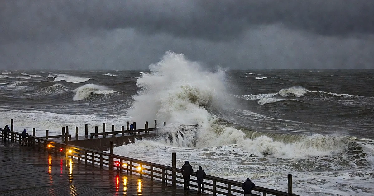 Massive storm waves crashing against coastal seawall with emergency lights reflecting on wet boardwalk at dusk