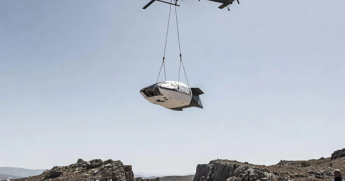 Aerospace test vehicle suspended from helicopter above Mediterranean terrain during high-altitude drop test