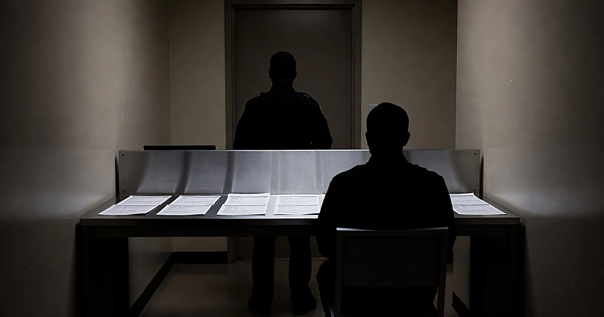 Federal booking station with silhouetted agents processing intake forms at metal desk under fluorescent lighting