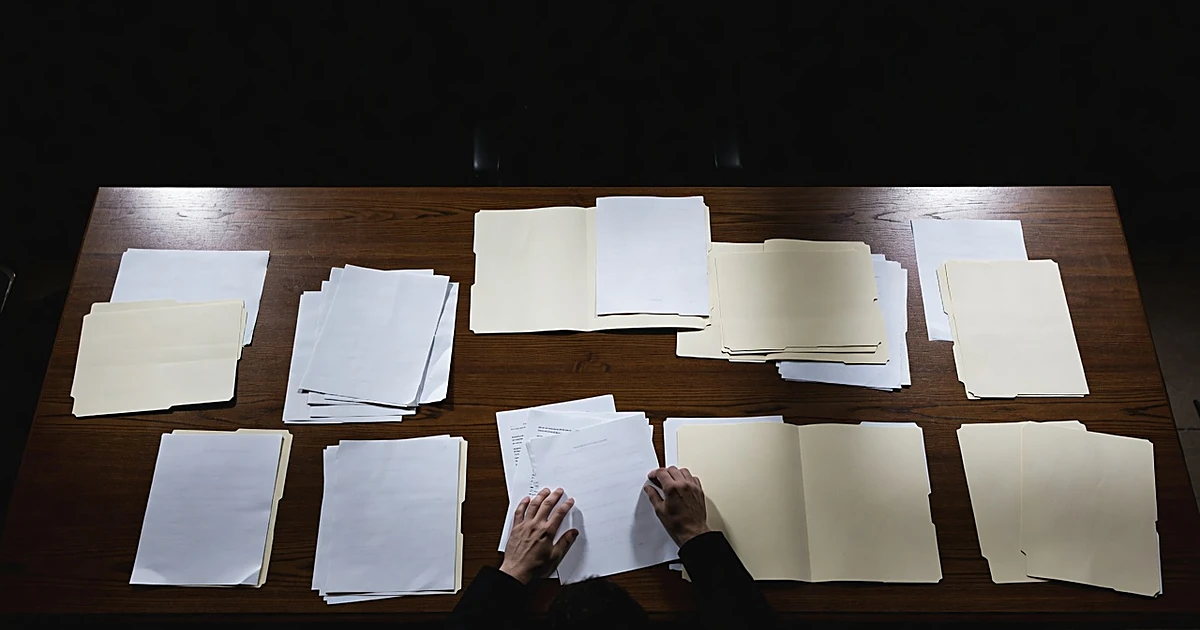 Documents and folders spread on evidence table with investigator's hands reaching toward papers