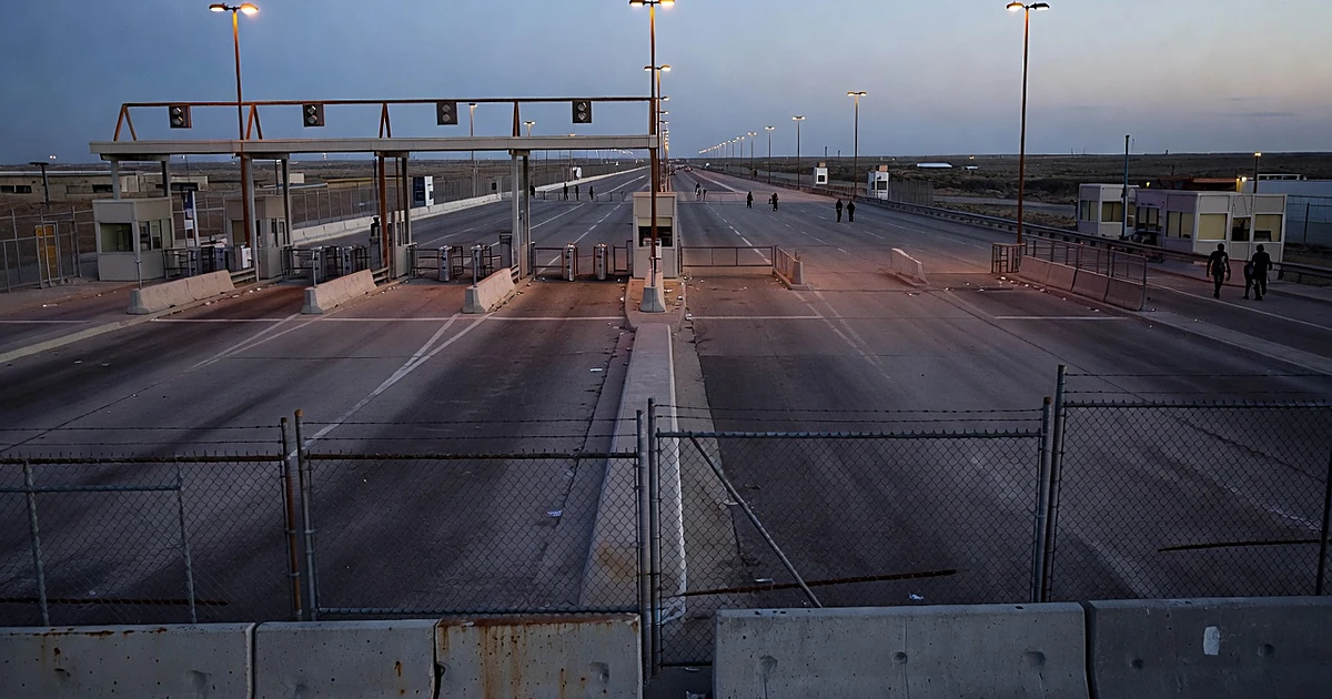 Empty border crossing lanes at dusk with overhead lights and security fencing stretching into the distance