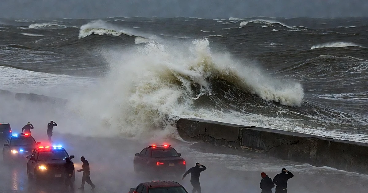 Massive waves crash against seawall as emergency vehicles with flashing lights respond to approaching hurricane conditions
