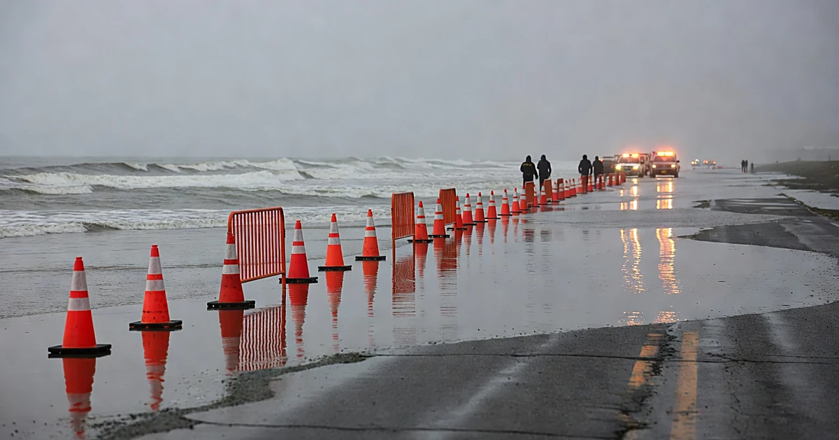 Flooded coastal street with emergency barricades and distant silhouettes of response personnel near vehicles