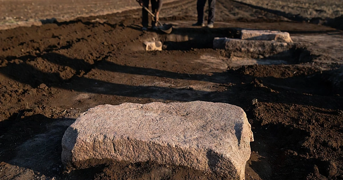 Pink granite block partially excavated from dark soil in agricultural field with silhouetted figures in background