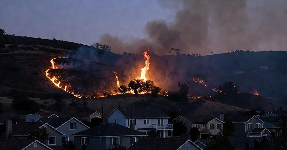 Wildfire flames burning through hillside brush near residential homes at dusk in Southern California terrain