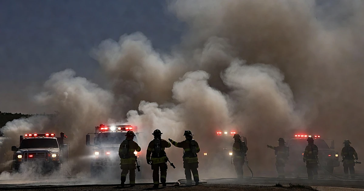 Wildfire flames consume hillside brush above residential homes as emergency lights flash and firefighter silhouettes respond
