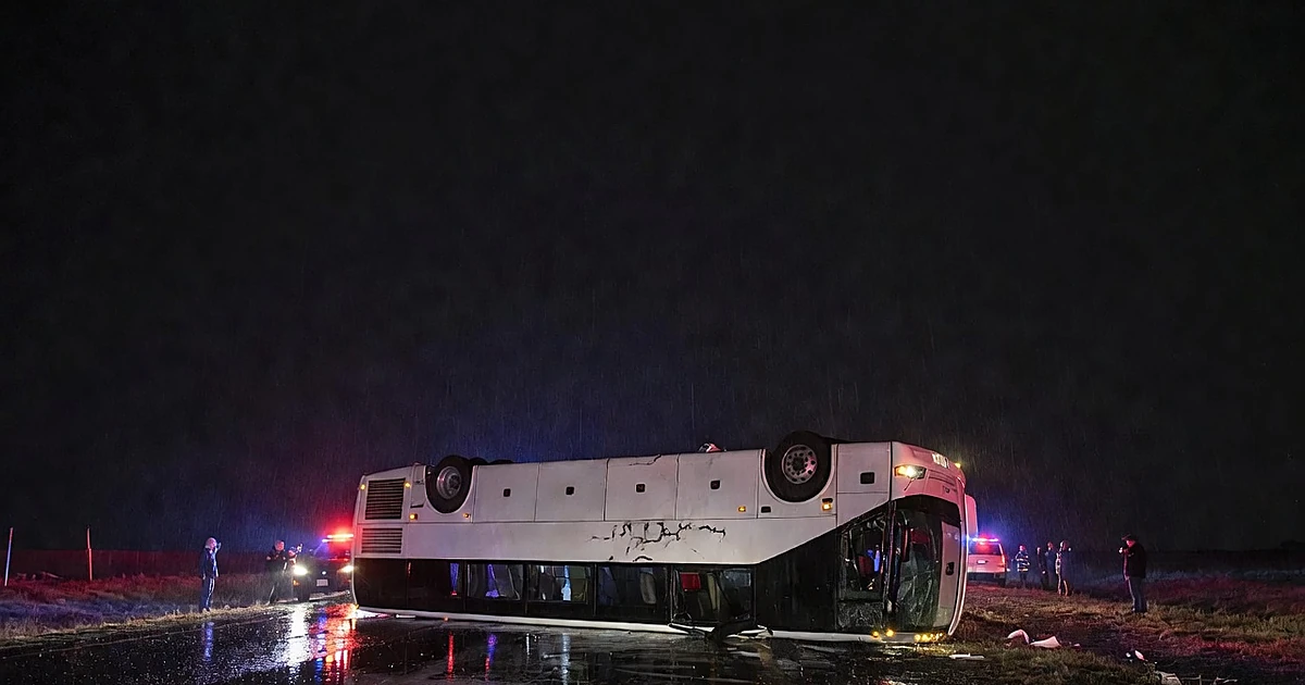 Overturned tour bus on highway shoulder at night illuminated by emergency vehicle lights in the rain