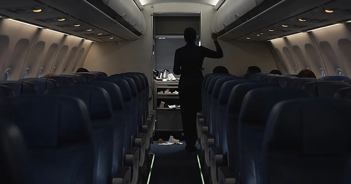 Airplane cabin interior with silhouetted flight attendant in galley area, scattered service items, emergency lighting