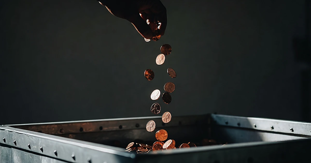 Silhouetted hand releasing cascade of pennies into industrial collection bin with dramatic lighting