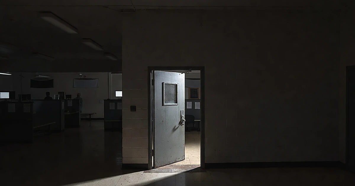 Police station booking area with heavy metal door partially open, cold fluorescent lighting on worn floor