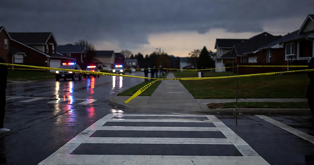 Police tape across wet suburban street at dusk with emergency lights reflecting on pavement