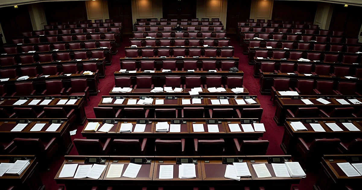 Empty legislative chamber with scattered papers on desks and vacant seats under warm overhead lighting