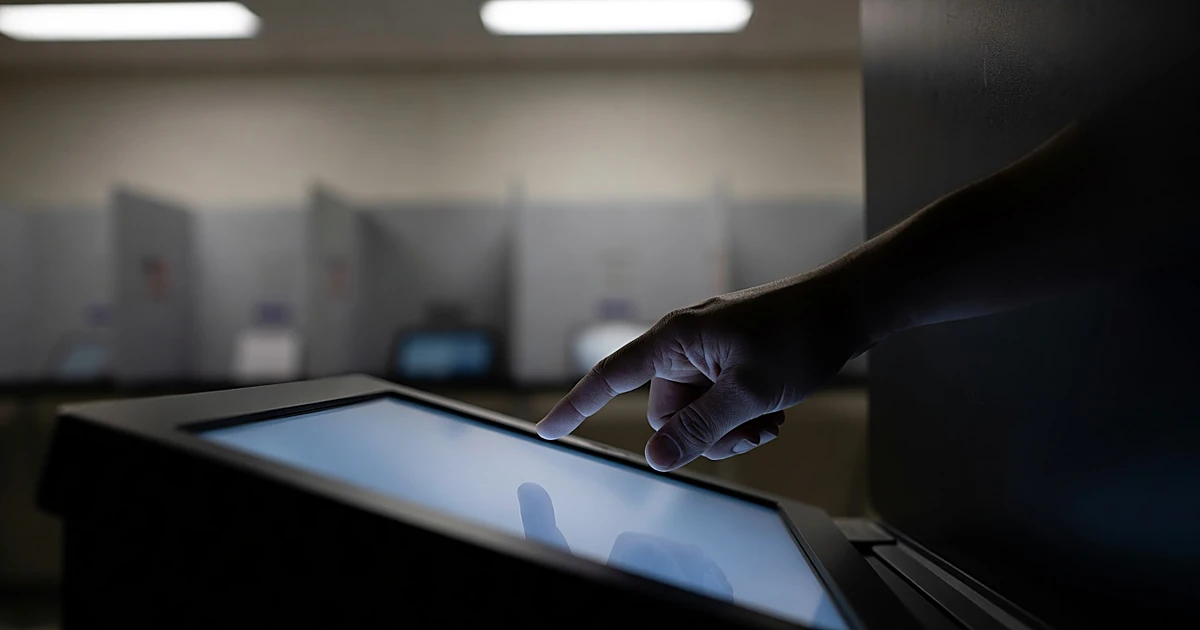 Silhouetted hand hovering over illuminated touchscreen voting machine in polling booth