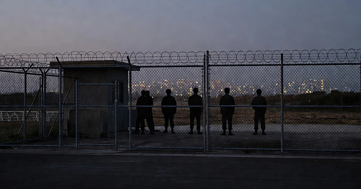 Military base security gate at dusk with silhouetted personnel and city lights beyond perimeter fence