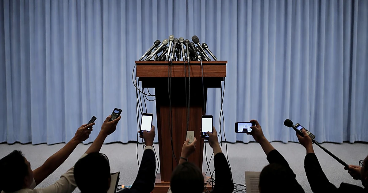 Empty White House press briefing podium surrounded by reporters' silhouetted arms holding recording devices forward